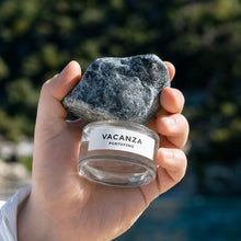 A hand holds a perfume bottle with label 'VACANZA PORTOFINO' with a dark gray stone resting on top, against a blurred outdoor background of greenery and water.