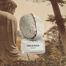 A clear glass perfume bottle labeled 'VACANZA KYOTO' sits on a white stone orb, set against a sepia-toned collage background featuring a large Buddha statue and trees.