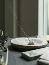 A thin incense stick stands in a small white incense holder on a round stone incense burner, set on a dark tabletop with dried flowers and books nearby in a softly lit indoor setting.