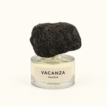 A clear glass perfume bottle with a pale yellow liquid and a cream label reading 'VACANZA MEGÈVE', topped with a large black porous stone cap, on a plain white background.