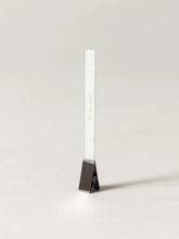 A slim white rectangular card or strip with gold vertical text, standing upright in a small dark metal holder against a clean light gray background.