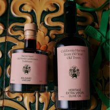 Two bottles of Heritage Old Trees balsamic vinegar and extra virgin olive oil displayed on a decorative green and gold tile surface with a green ribbon.