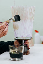 Person using a brush to mix contents in a glass container on a glass surface.