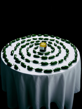 Round table with a white tablecloth, green peppers arranged in concentric circles, and a candle in the center.