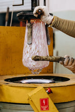Person working with a glassblowing tool on a pink glass object in front of a yellow machine.