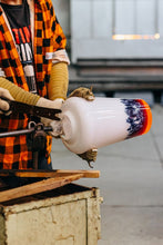 Person working with a glassblowing tool and a colorful glass piece in a workshop setting.