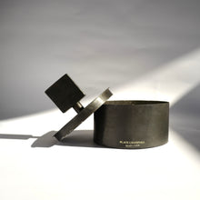 Black cylinder metal Mad et Len Bougie Totem Square Grande - Black Champaka with square totem lid handle resting on the side. 