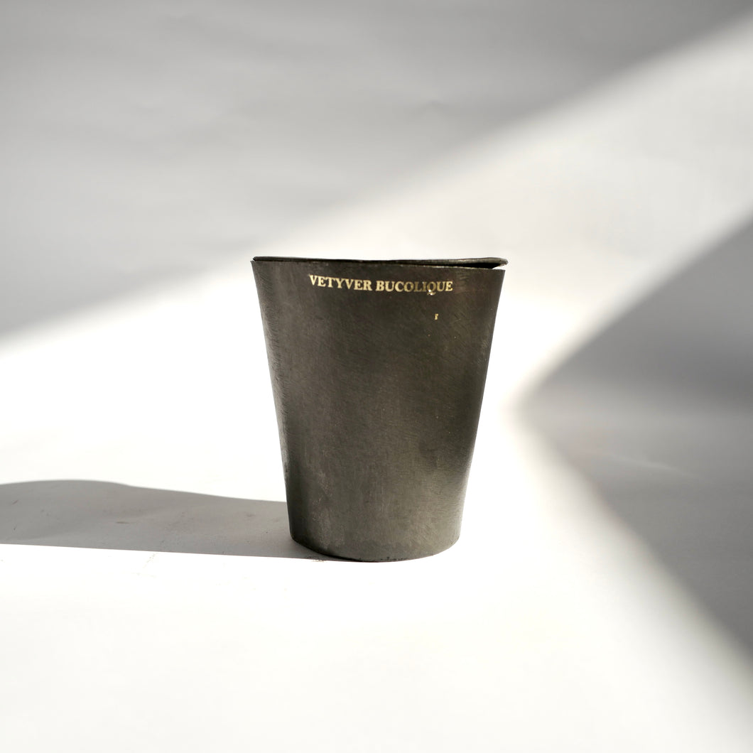 Black metal tapered Mad et Len Bougie Vestimentale Petite - VETYVER BUCOLIQUE (Black Wax) container with lid. Gold appliqué label of candle sent name. 