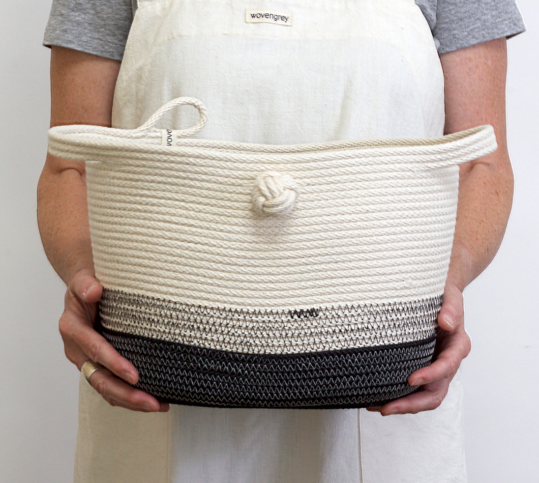 A person wearing an apron holding the Magazine Bucket with Knot. Black and white basket with sewing stitches. 