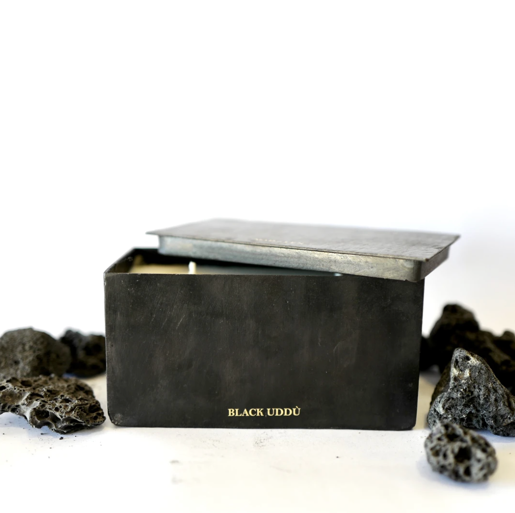 Black Rectangular shape metal candle container with lid opened ajar. Mad et Len Black Block Horizontal - BLACK UDDÙ candle. Lava rocks by the side of the candle. 