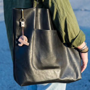 A person holding a leather back on shoulder. 1.61 Soft Goods Cactus Sun Key Fob (Key Hook) Nude color hanging from a black leather back. 